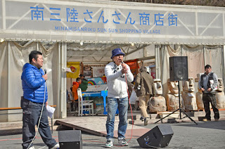 南三陸さんさん商店街の前のスペースでマイクを持った男性2人が並んで立っている写真