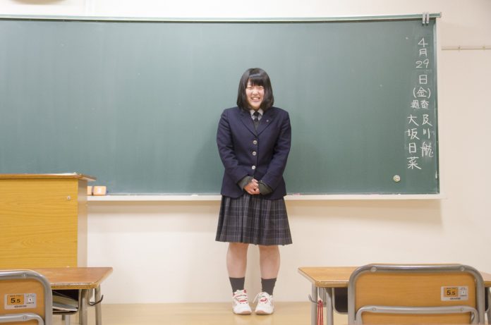 教室の黒板の前に立った制服姿の大坂さんが笑顔で写っている写真