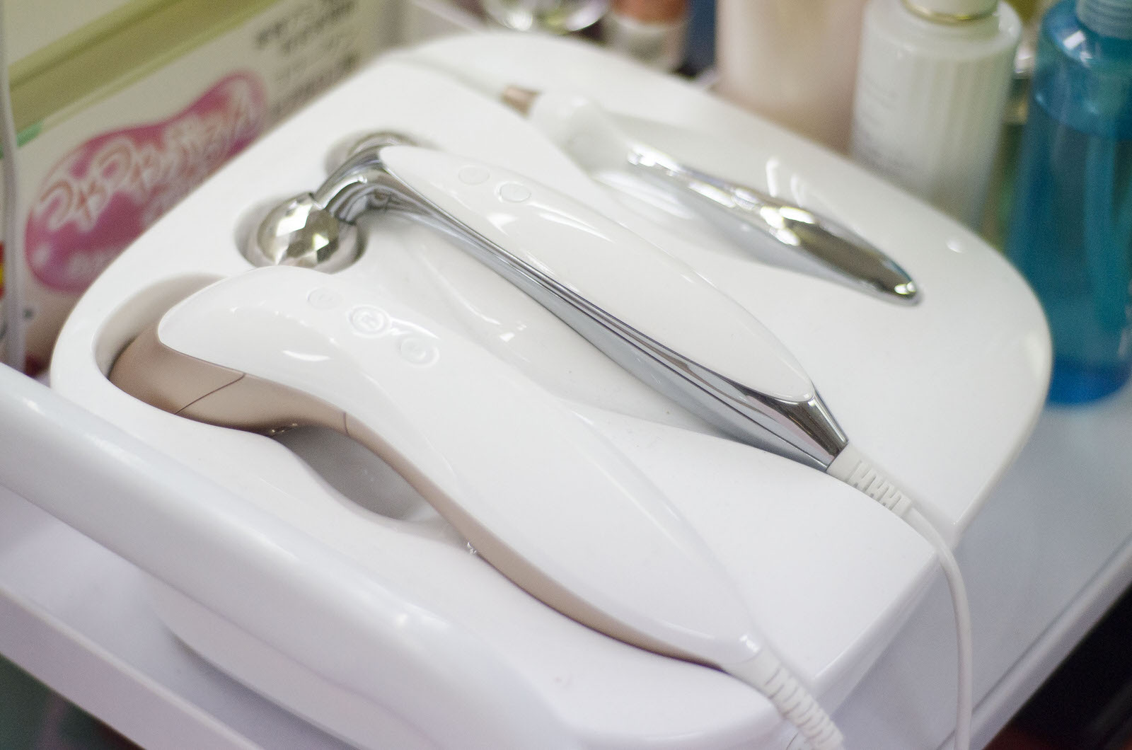 化粧品のボトルが置かれている白い台の上に、白と銀で構成されている数本の美容機器が整然と並べられている写真