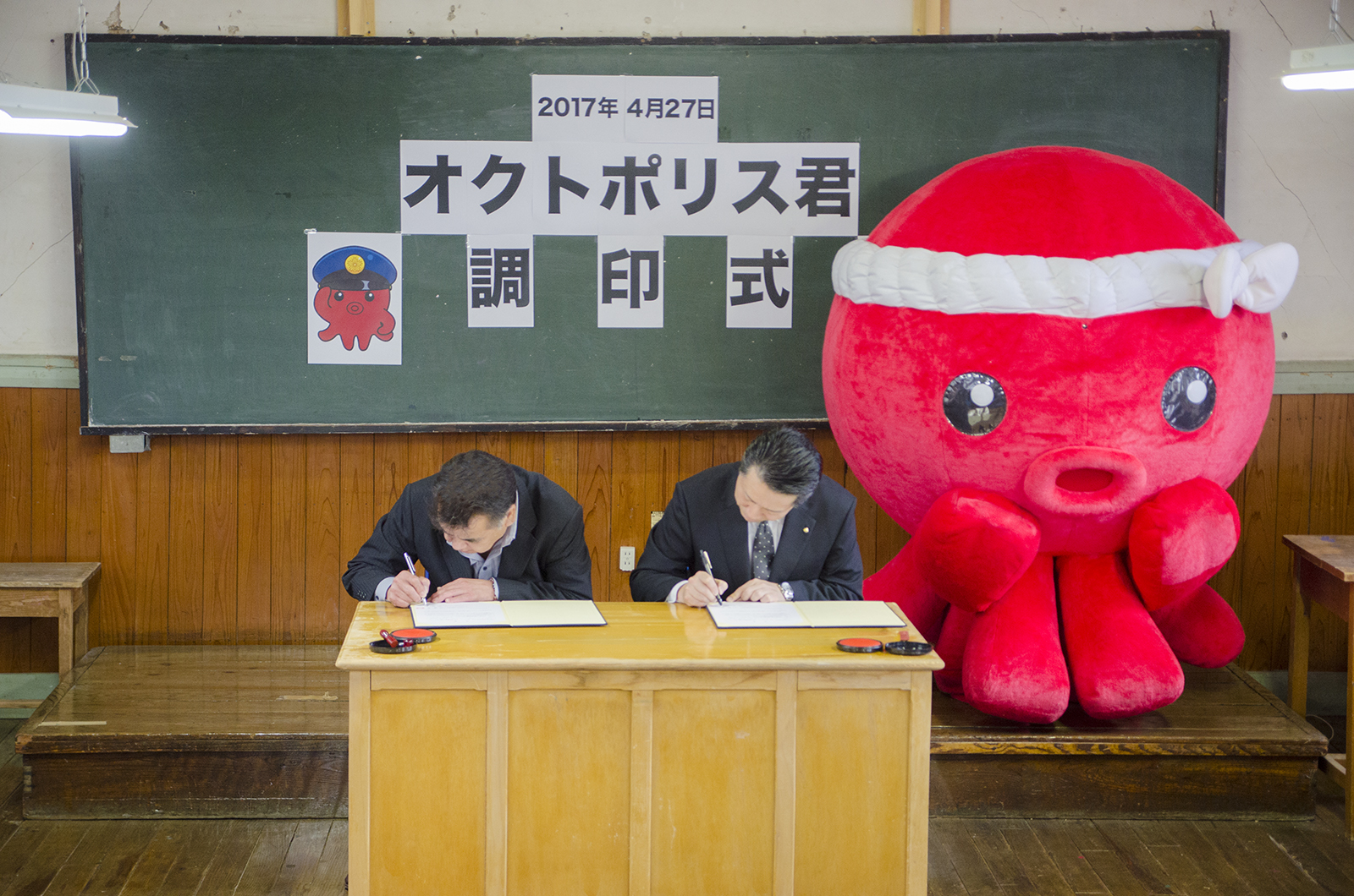 「2017年4月27日 オクトポリス君調印式」の文字とオクトポリス君のイラストがい掲示された黒板前にオクトパス君の着ぐるみが立ち、その前に置かれた机で佐藤さんと高橋さんが調印の署名を行っている様子の写真