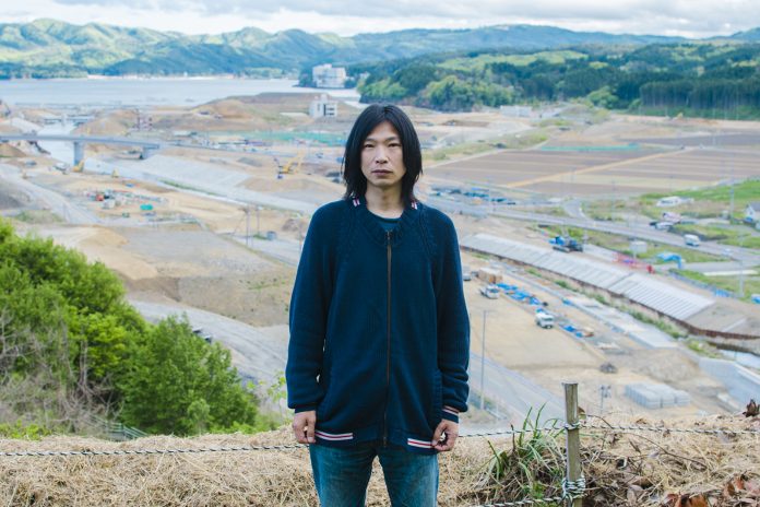 復興工事が進む広大な造成地と山並みを背景に、紺色の上着を着た佐藤さんが正面を見て立つ写真