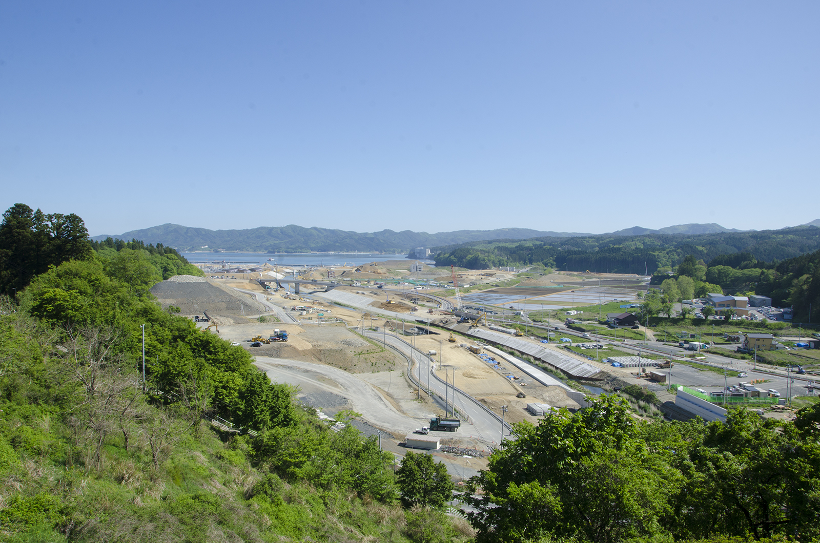 青空の下、山に囲まれた広大な土地で道路や橋などのインフラ整備が進められており、複数の重機が稼働する中、造成中の土地や建設資材が整然と配置された復興工事の様子が一望できる写真