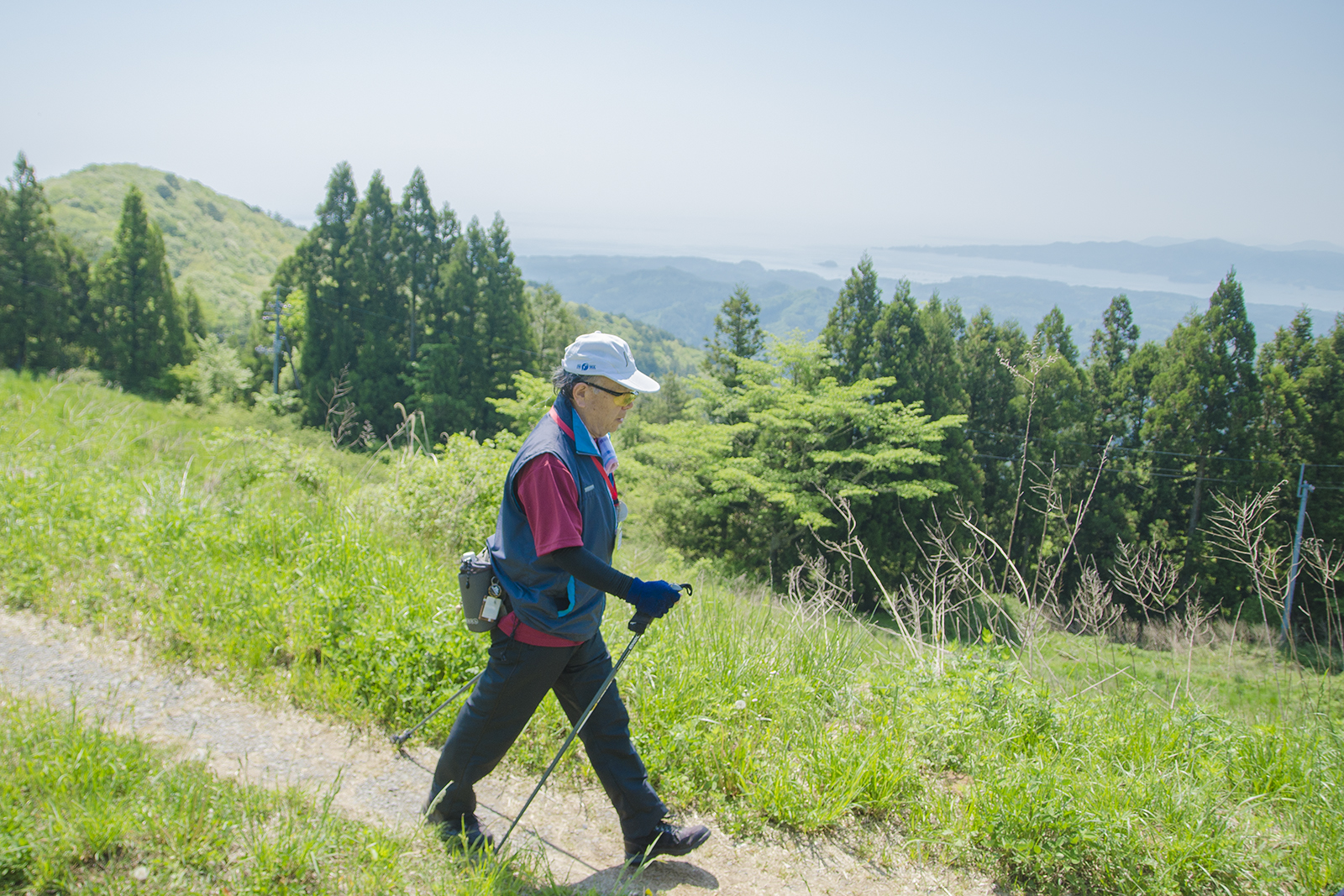 帽子をかぶった男性がスティックを持って、緑に囲まれた見晴らしの良い山のトレイルコースを歩いている写真