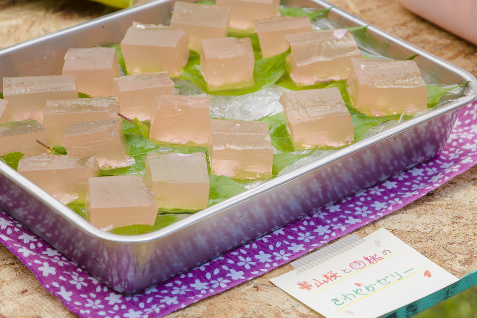 銀色のトレーに緑色の葉の上にピンク色の四角いゼリーを乗せた涼しげなお菓子がたくさん並べられ、前に「山桜と桃のさわやかゼリー」と書かれたメモが添えられている写真