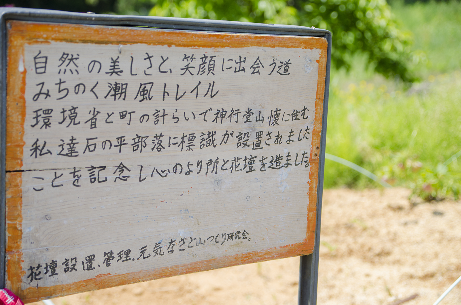 「自然の美しさと、笑顔に出会う道 みちのく潮風トレイル 環境省と町の計らいで神行堂山懐に住む私達石の平部落に標識が設置されましたことを記念し心のより所と花壇を造ました。花壇設置、管理、元気なさと山つくり研究会。」と手書きで書かれた案内板が立っている写真