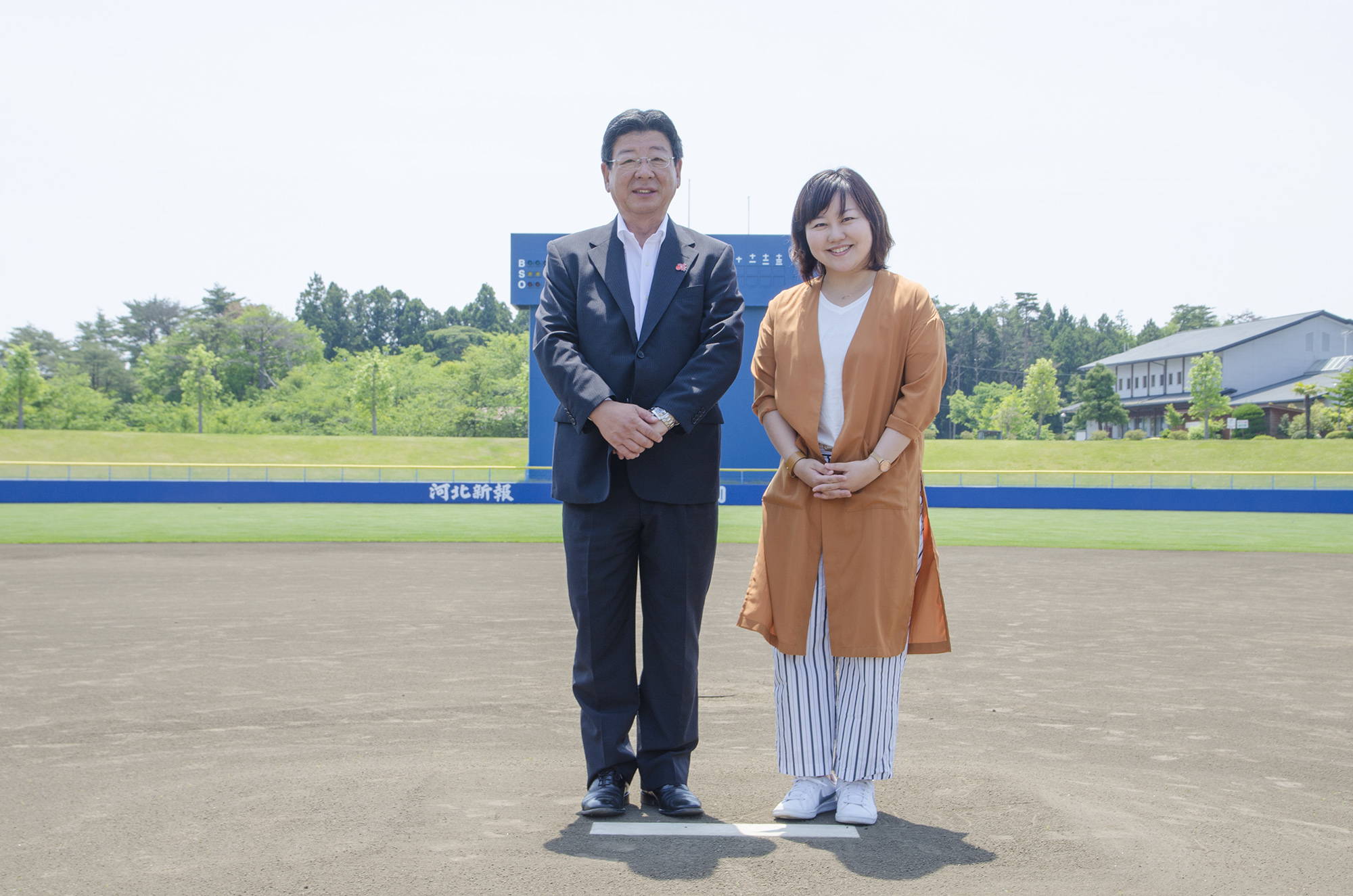 青いスコアボードの後ろに緑の森が広がる野球場のホームベースに佐藤仁町長と理解の女性が並んで立っている写真