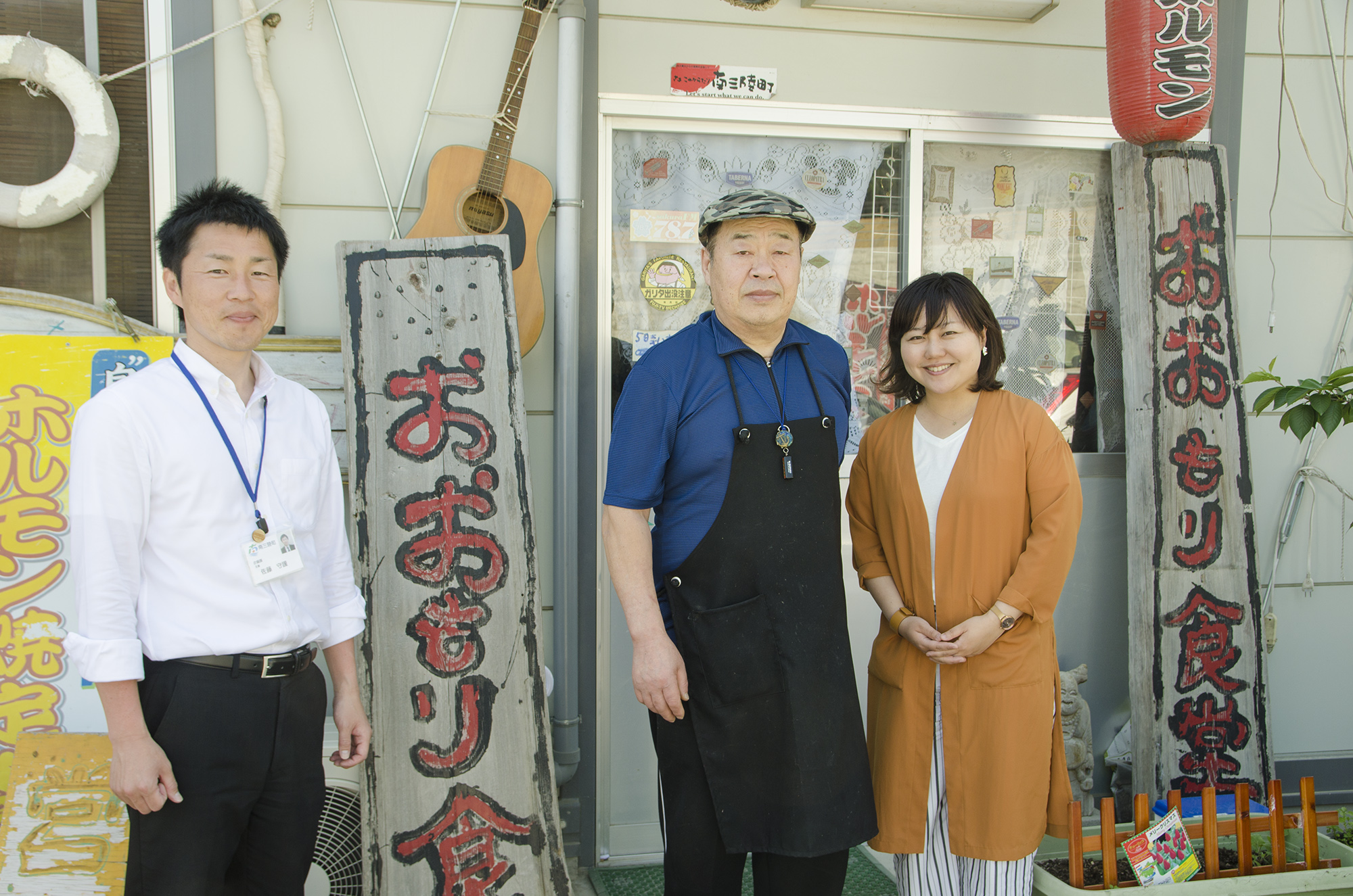 白シャツ姿の男性、黒いエプロンを着けた男性、リポーターの女性との3人が、赤い提灯と木の看板が掲げられた食堂の入口前で並んで立っている写真