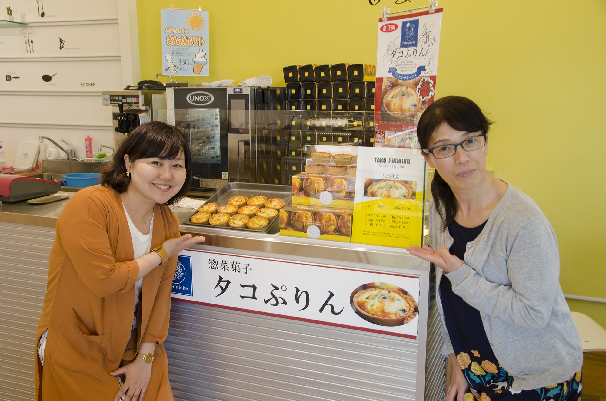 「惣菜菓子タコぷりん」と書かれたカウンターの上に商品が並んでおり、その両側で商品を手で示している女性リポーターと店の女性の写真