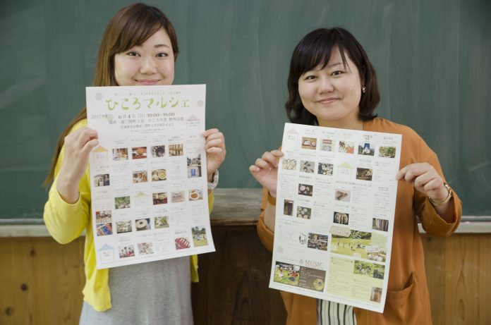 黒板が設置された壁の前で、ひころマルシェのチラシを持った中島さんと女性リポーターが並んで立っている写真