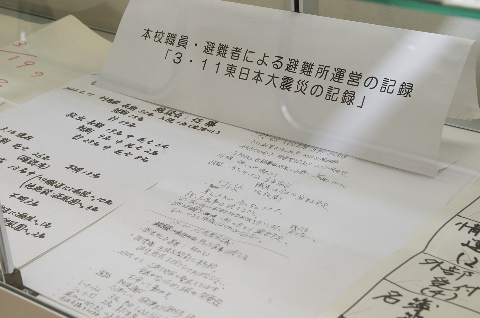 「本校職員・避難者による避難所運営の記録「3・11東日本大震災の記録」」と書かれた札の前に手書きで記録された資料が並んでいる写真