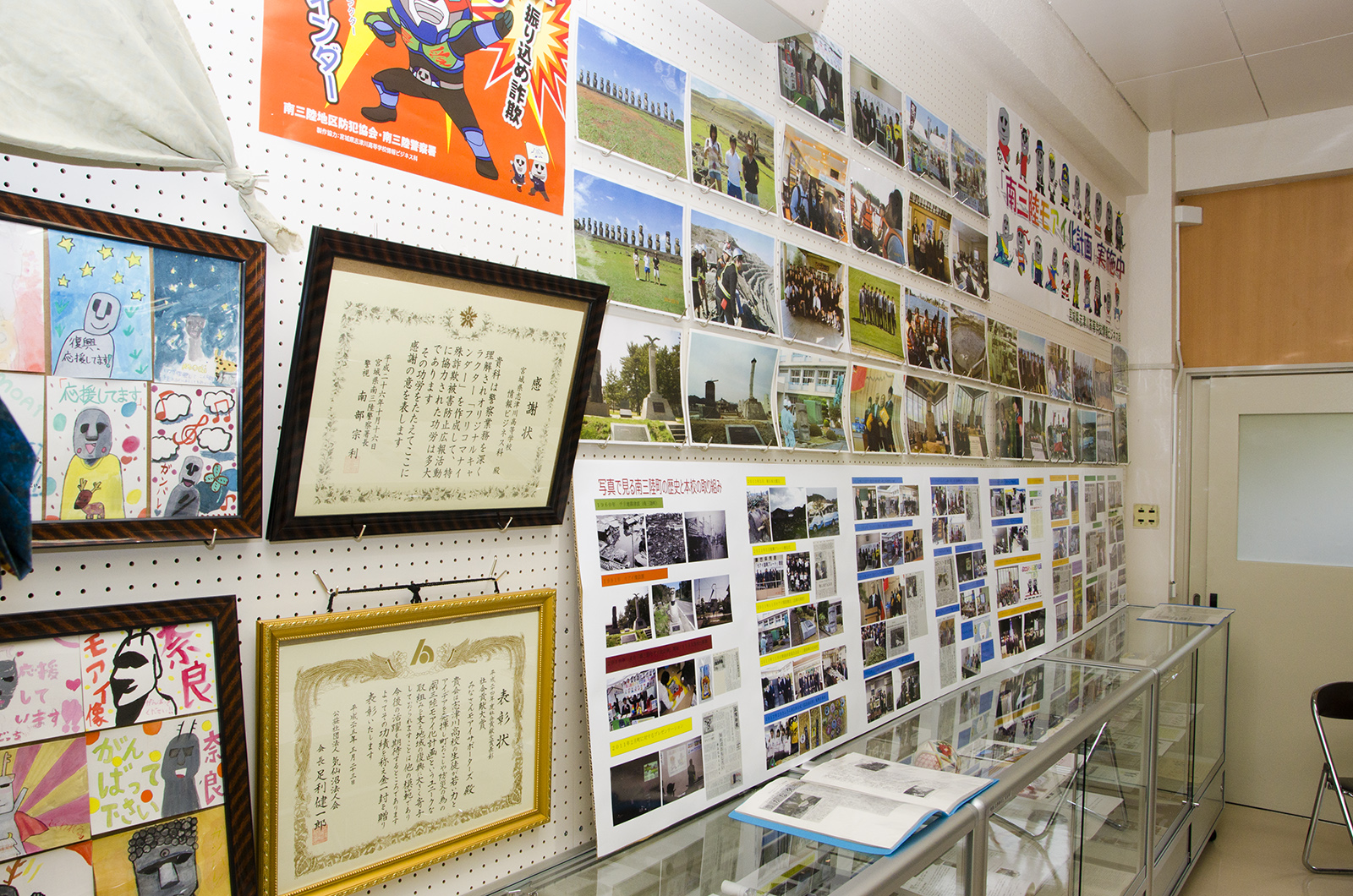 イラストや賞状、たくさんの写真が掲示された壁の下に、資料が入ったガラスの展示ケースが設置されている写真