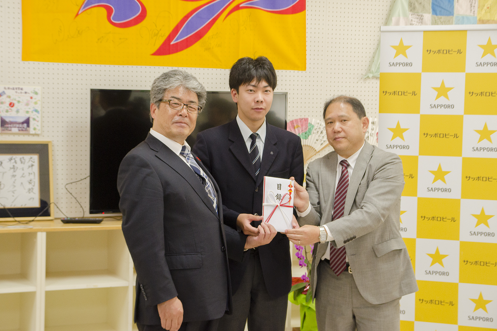 サッポロビールのバックパネルや大きなモニターの前でスーツ姿の3人の男性が目録の封筒に手を携えて立っている記念写真