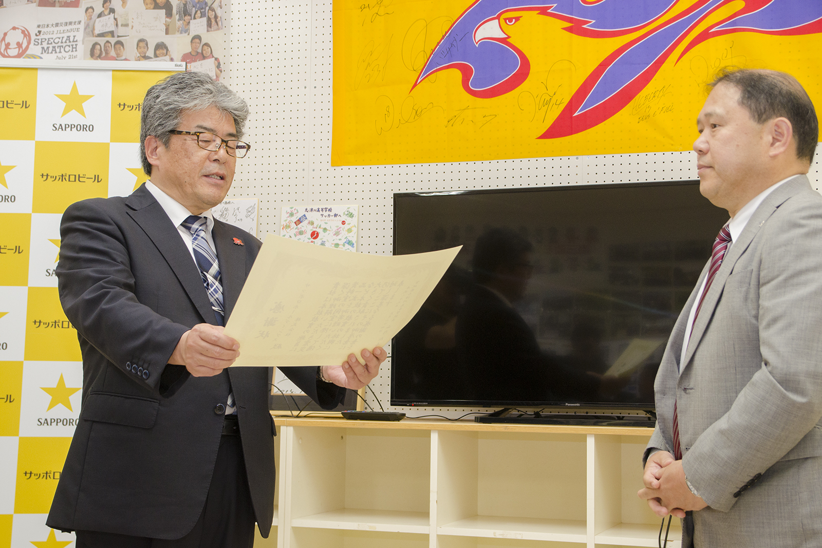 大きなモニターが設置された棚の前で、志津川高等学校校長山内松吾さんが向かい合って立った梅里俊彦さんに感謝状を読み上げている写真