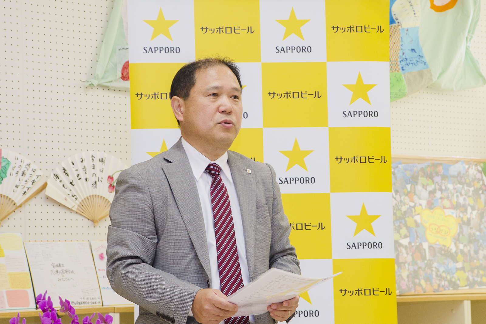 サッポロビールのバックパネルの前で、資料を手に持ったサッポロホールディングス株式会社コーポレートコミュニケーション部部長梅里俊彦さんが話をしている写真