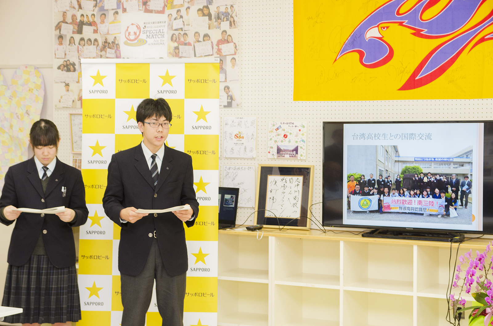 写真や色紙などが掲示され、サッポロビールのバックパネルが置かれた壁を背に制服姿の男女2人の生徒が発表を行っており、棚の上のモニターに資料が映し出されている様子の写真