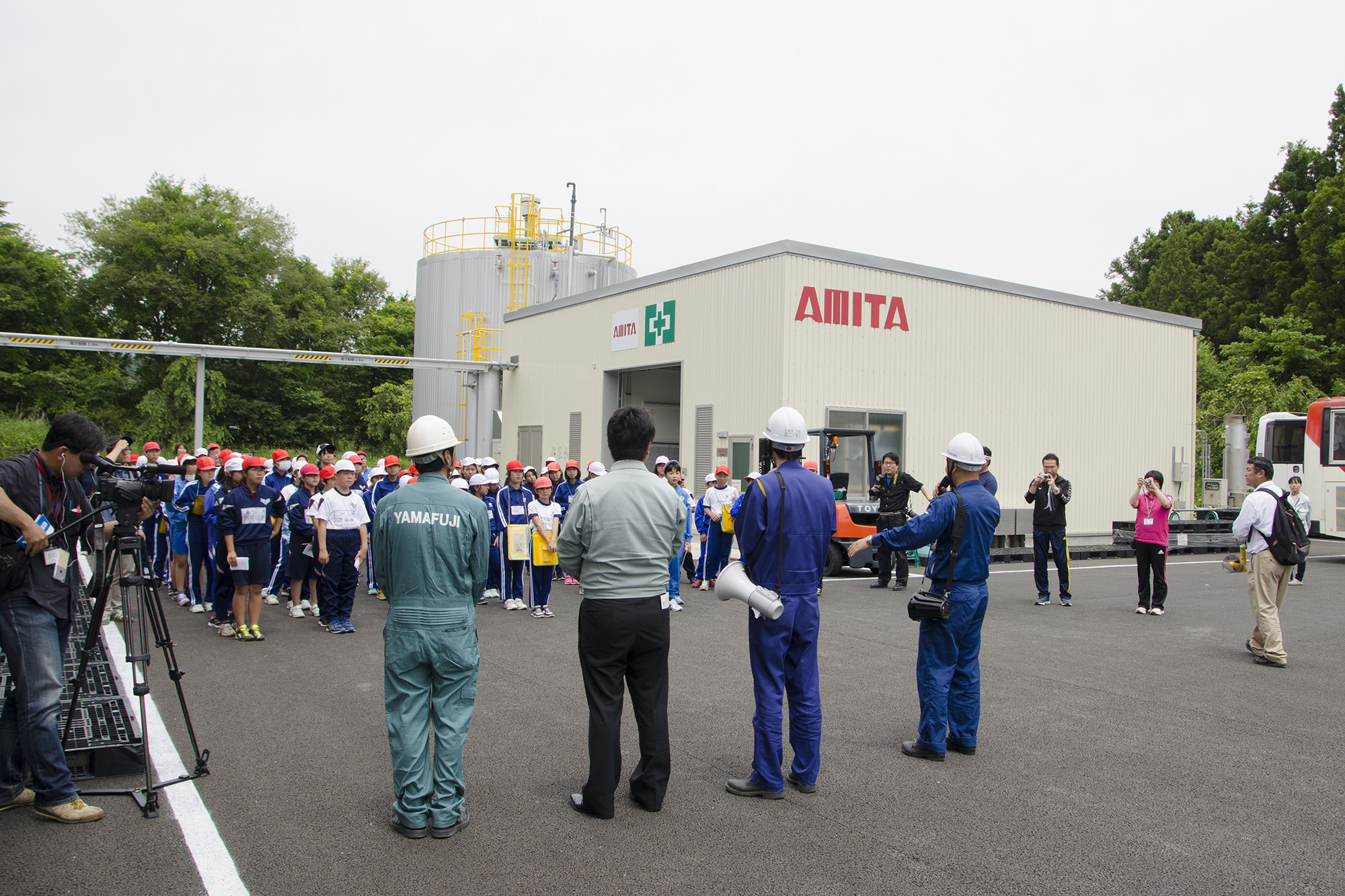 バイオガス施設の「AMITA」と書かれた建物前の広場に、赤い帽子をかぶった多くの小学生が整列し、作業服とヘルメット姿の工場関係者が拡声器を持って説明をしている様子を写した写真
