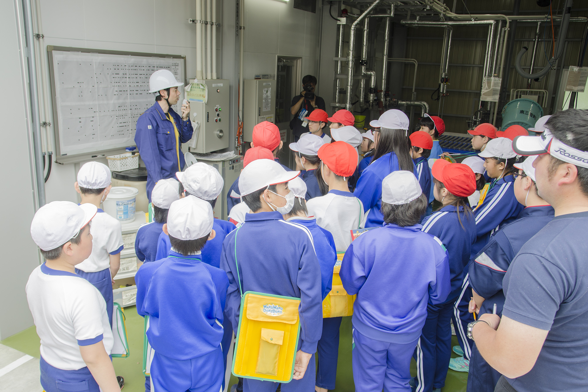 バイオガス施設の室内で、ヘルメットをかぶった作業員が操作盤の前で拡声器を使い、赤白の帽子をかぶった小学生たちに設備の説明をしており、子どもたちは真剣に話を聞いている写真