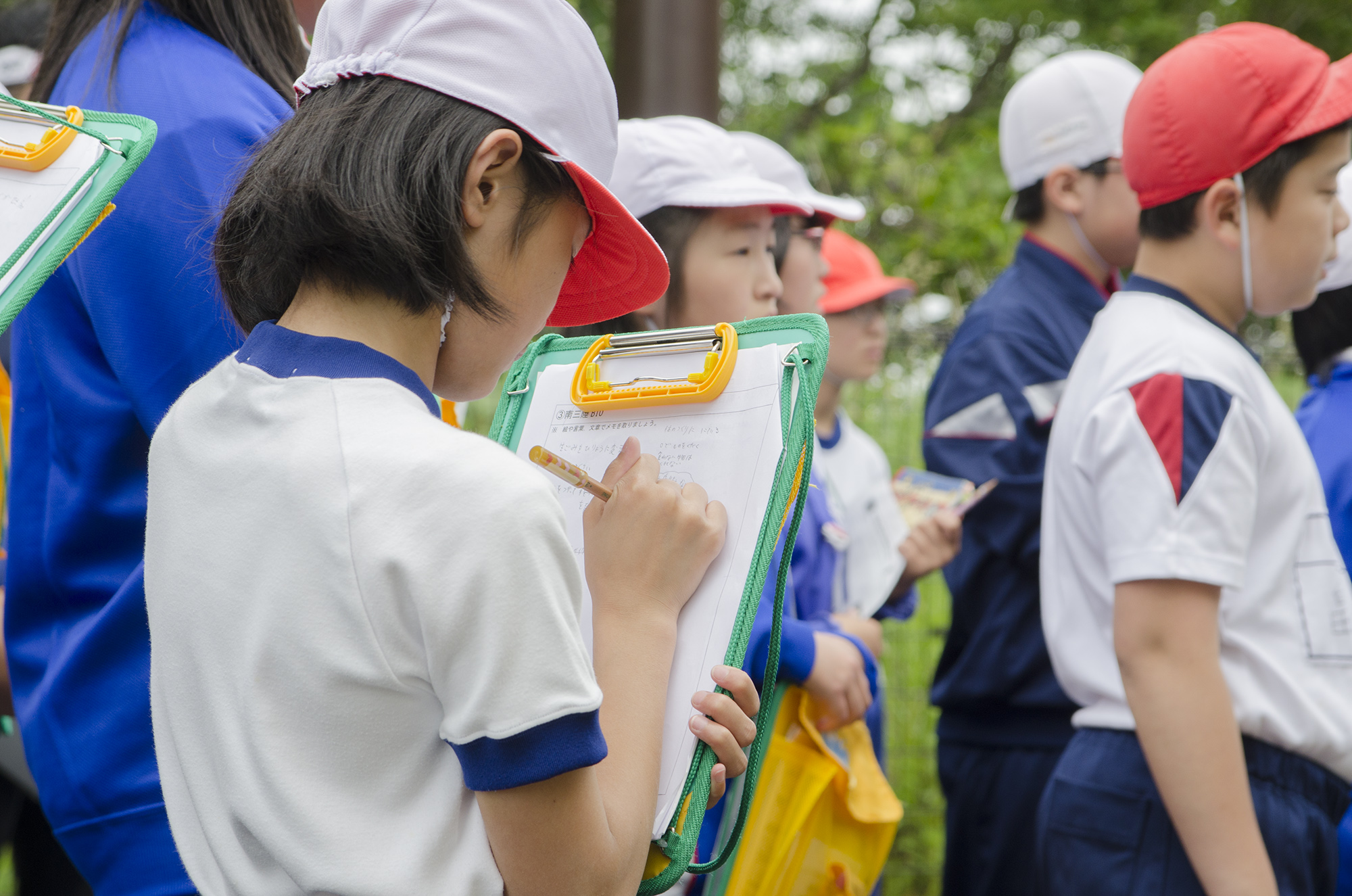 体操服を着て白い帽子をかぶった女子生徒がクリップボードを持ち、一生懸命メモを取っている様子を写した写真