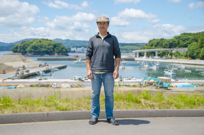 船の停泊する静かな漁港や緑の山々、高架道路が見える風景をバックに立っている佐々木昌則さんの写真