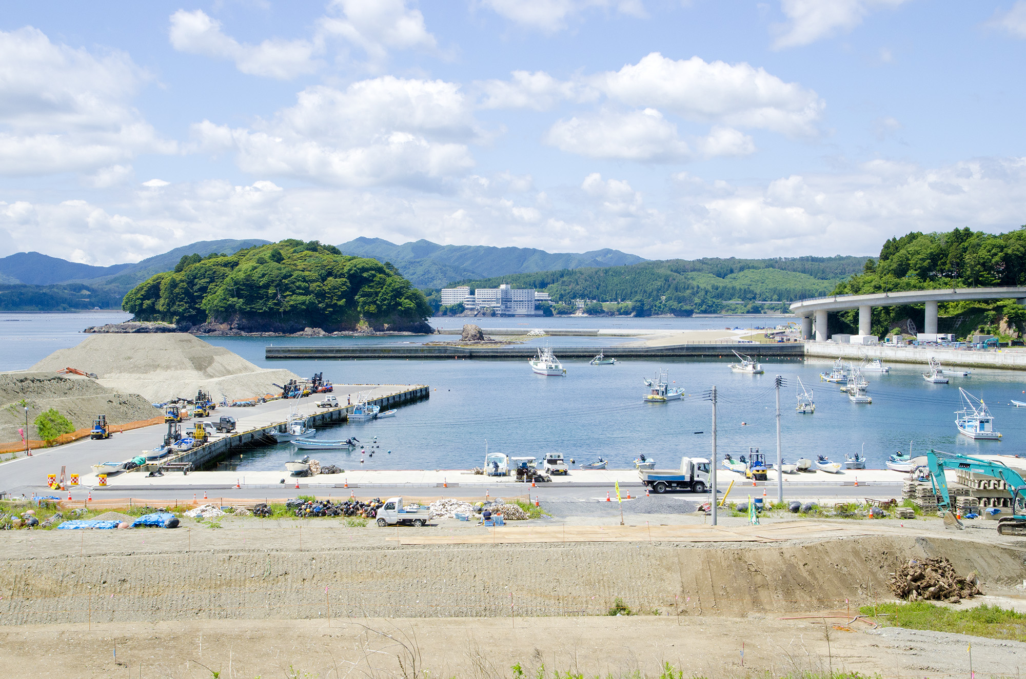 多くの漁船が停泊している港を中心に、奥には緑に覆われた小島と静かな海、さらにその向こうには山並みと建物が見える風景の写真