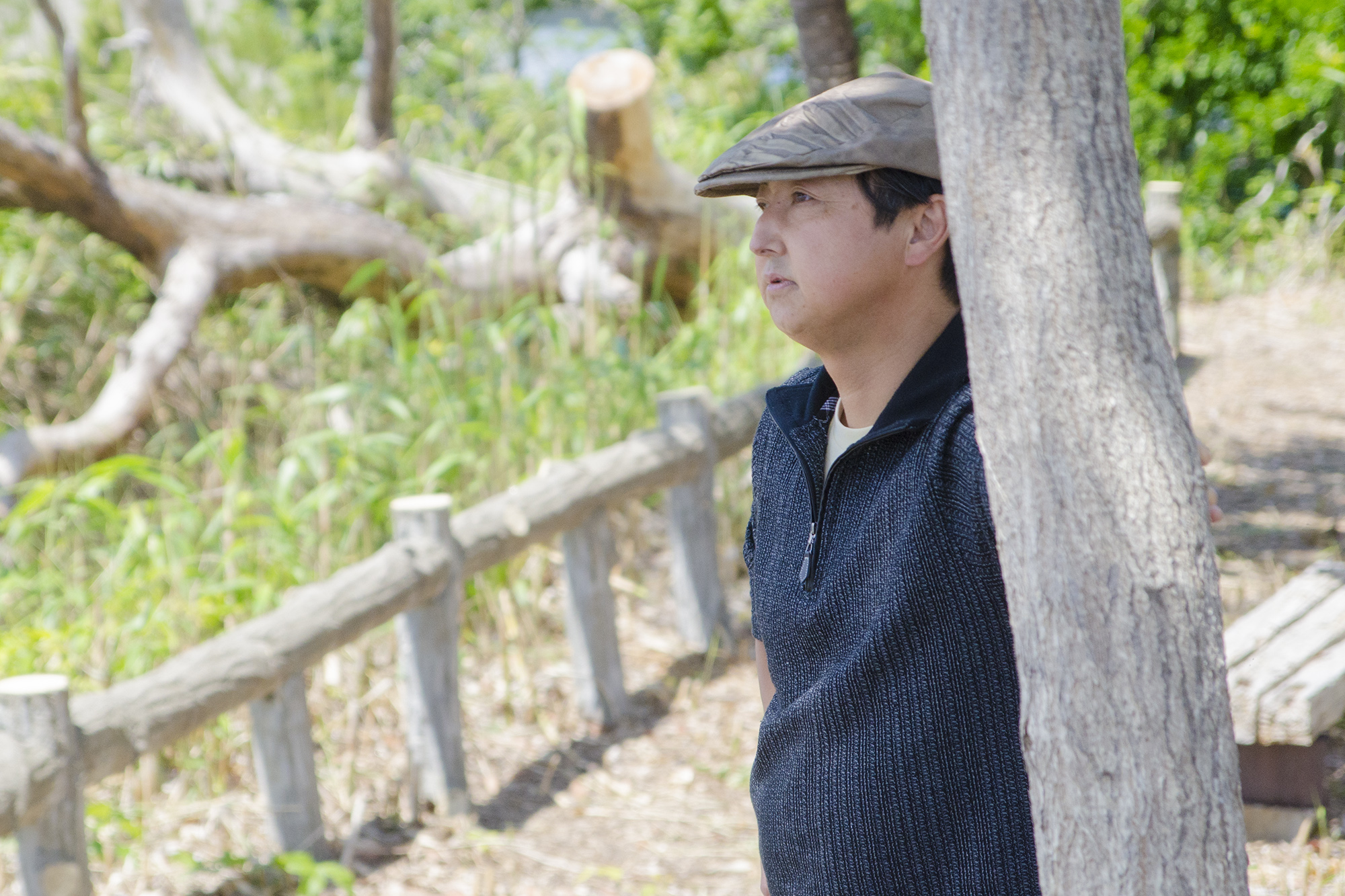 木製の柵や切り株、草木が広がる自然豊かな遊歩道の一角の木陰で、帽子をかぶった佐々木さんが、静かに前方を見つめながら立っている写真