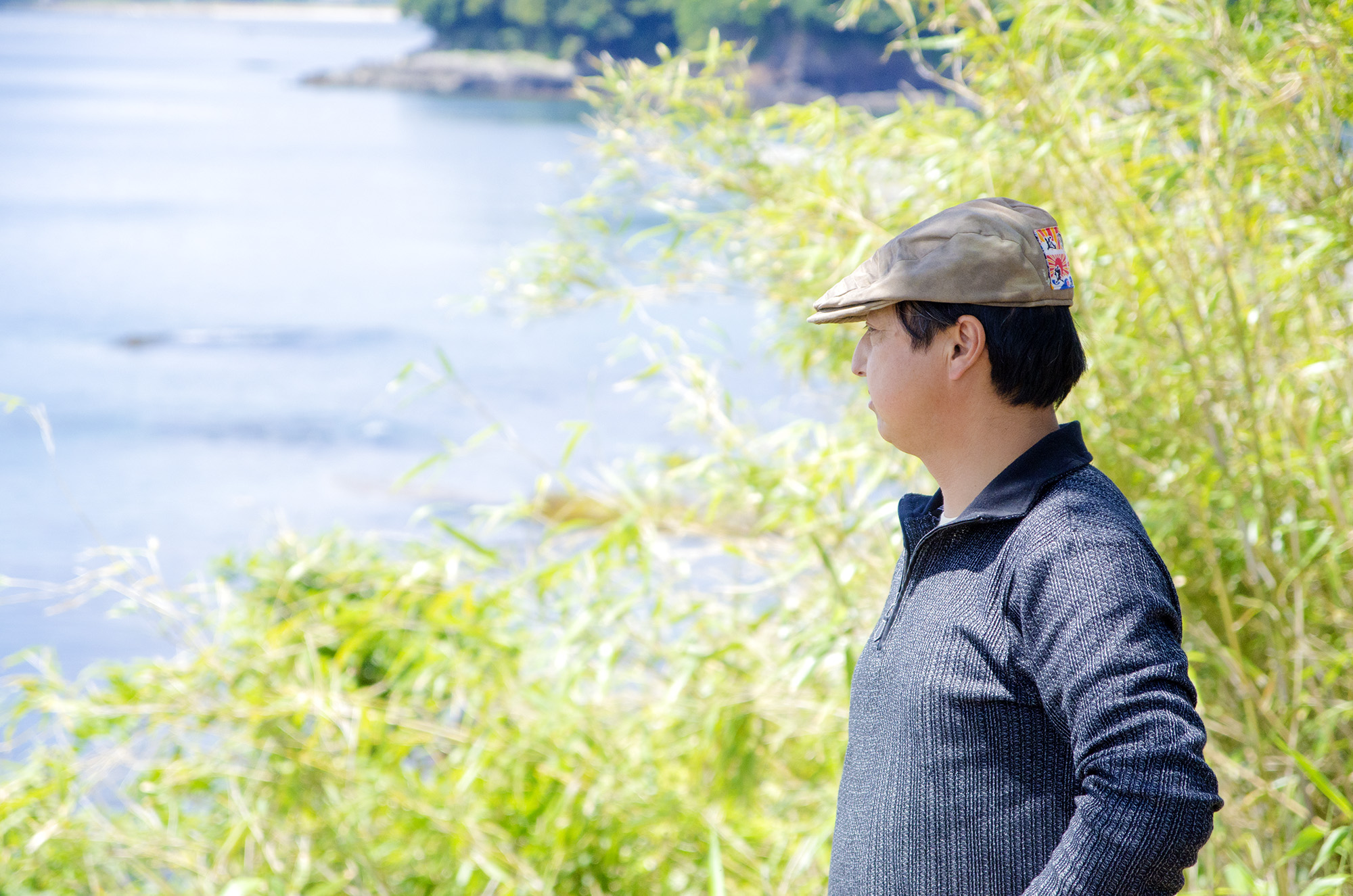 帽子をかぶった佐々木さんが、水辺に面した緑に囲まれた場所で穏やかな表情で遠くを見つめている写真