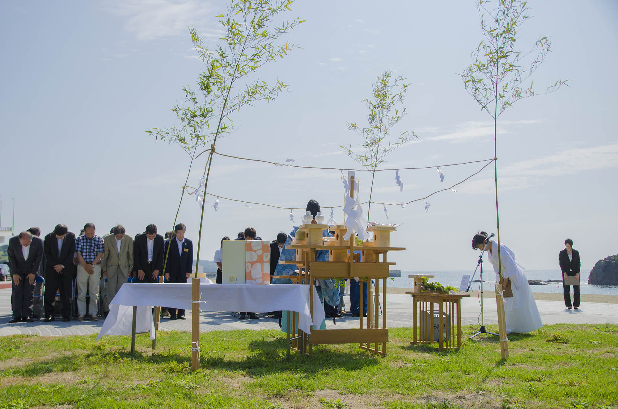 竹の柱で囲まれた祭壇の前で、神職と数名の参列者が頭を下げて祈りを捧げている、海岸沿いで行われている安全を祈願を祈祷している写真