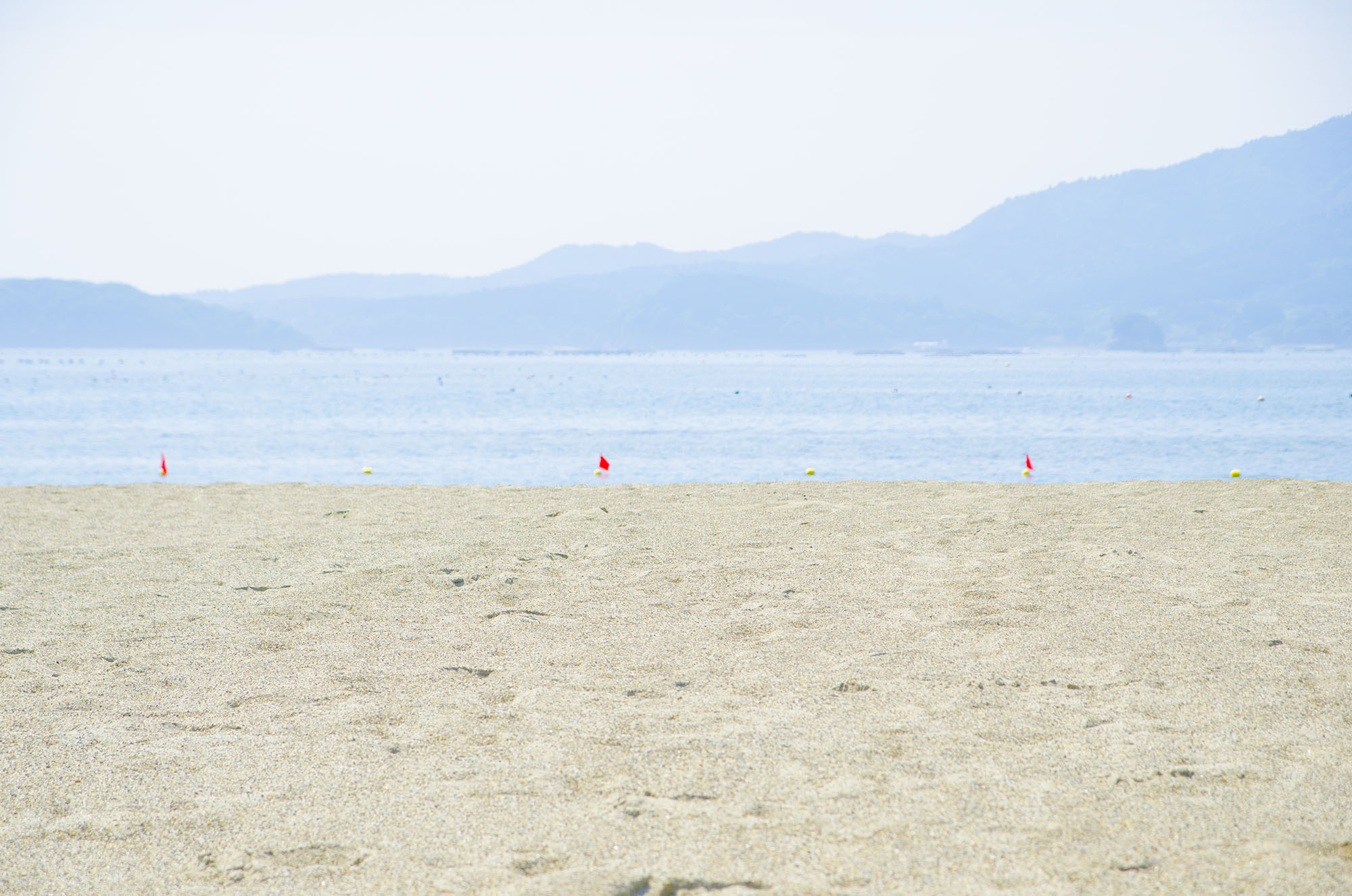 広くて平らな砂浜が広がり、その奥に穏やかな海と遠くの青い山々が見える、晴れた日の静かな海岸の写真