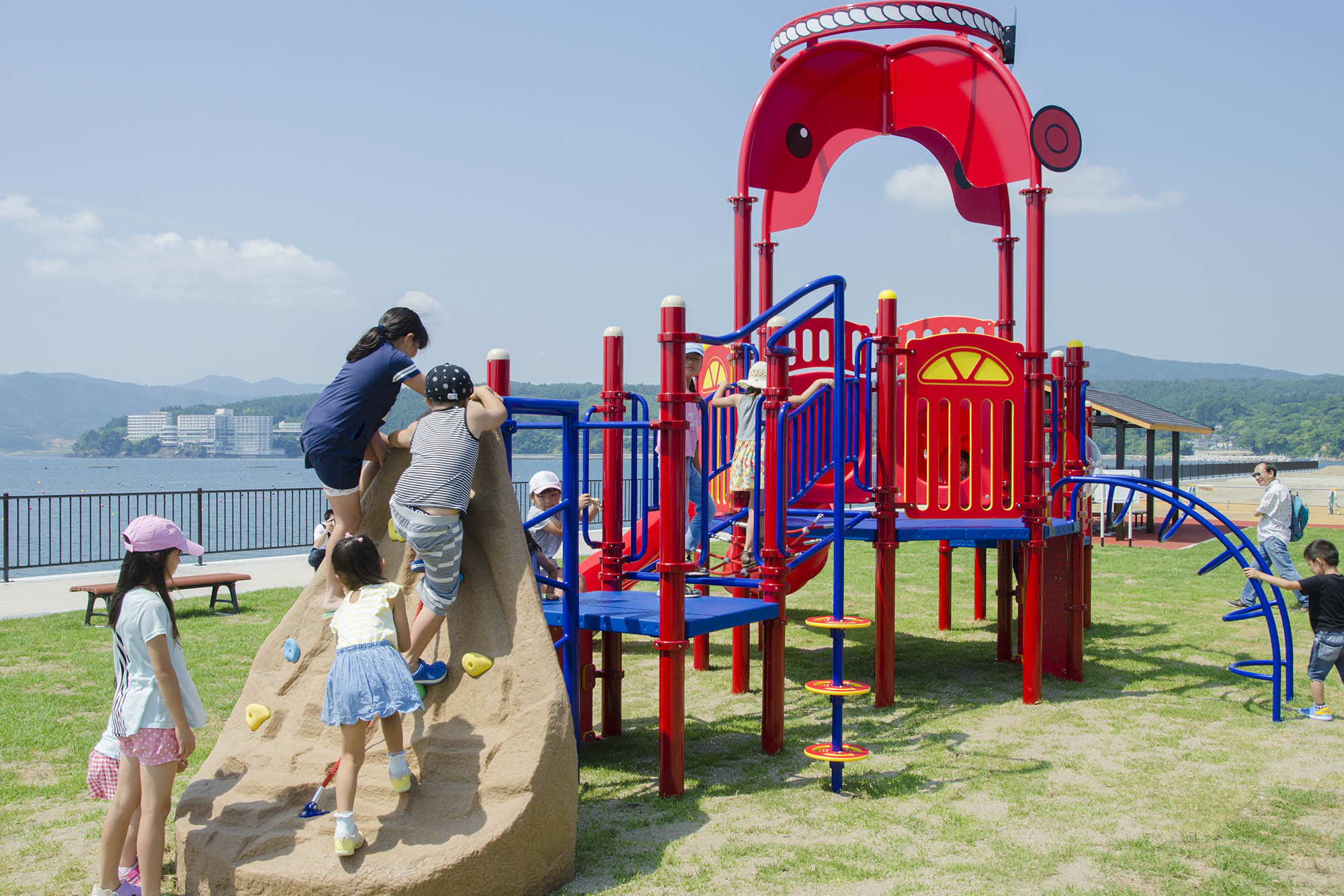 海辺にある公園で、複数の子供たちがオクトパス君をモチーフにした赤色の遊具を使って元気に遊んでいる写真