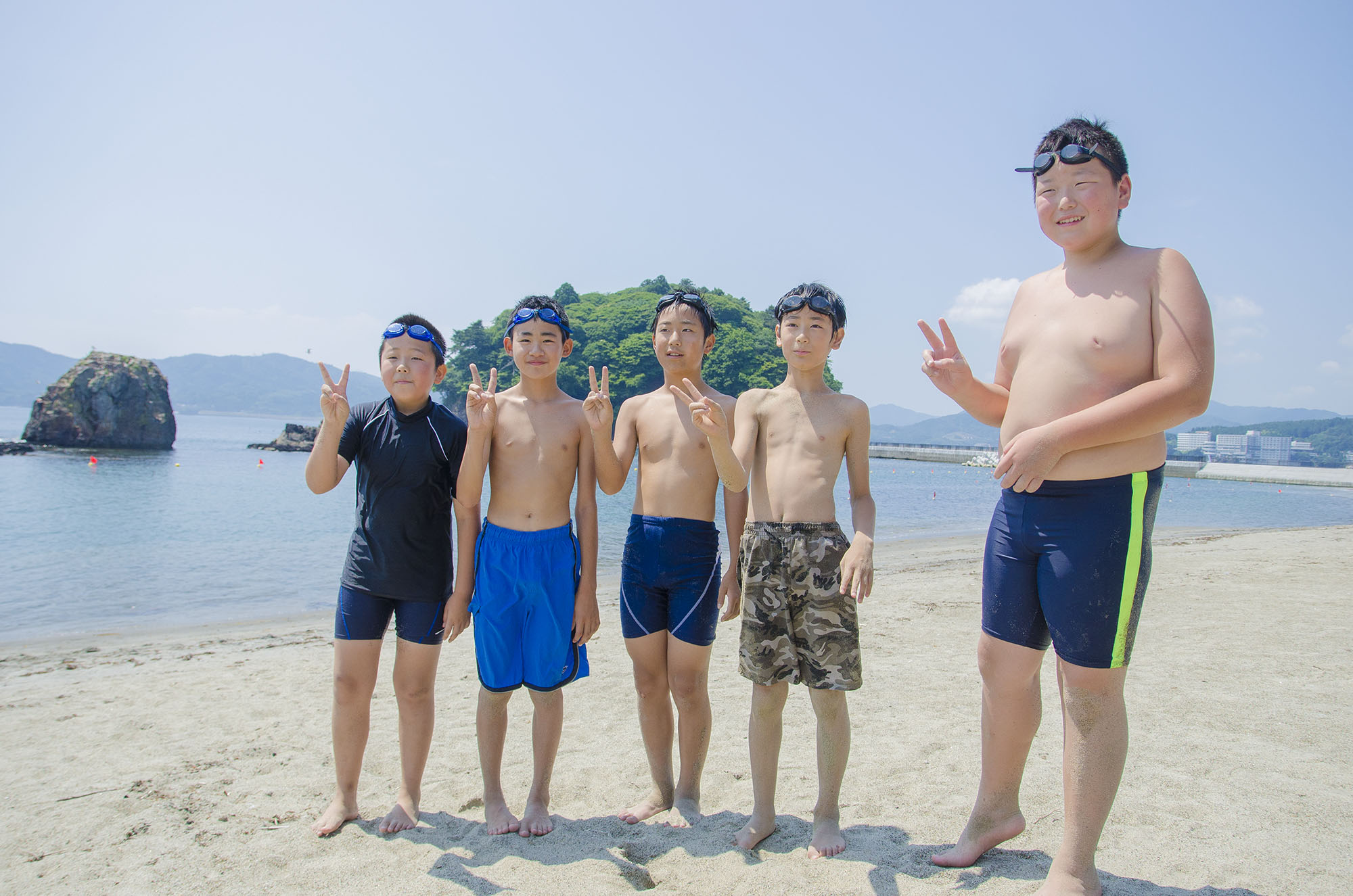 海辺の砂浜で水着を着た5人の小学6年生の男子児童が横一列に並び、笑顔でピースサインをしている写真