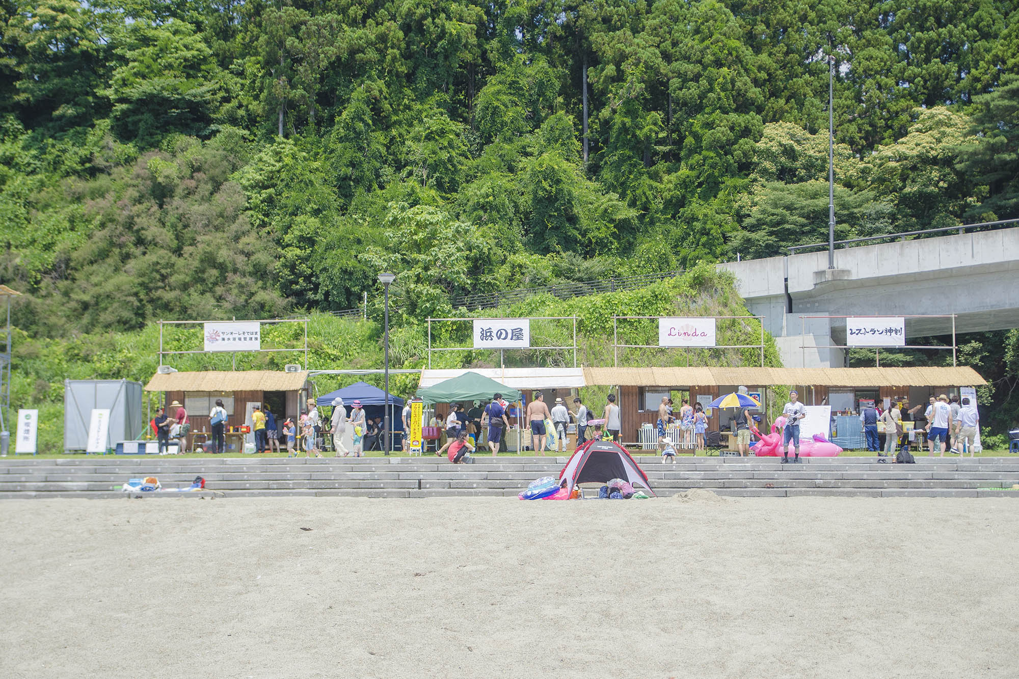 緑の木々を背景にして、砂浜の手前に複数の屋台が並び、その前に多くの人々が集まっている様子を写した夏の海辺の写真