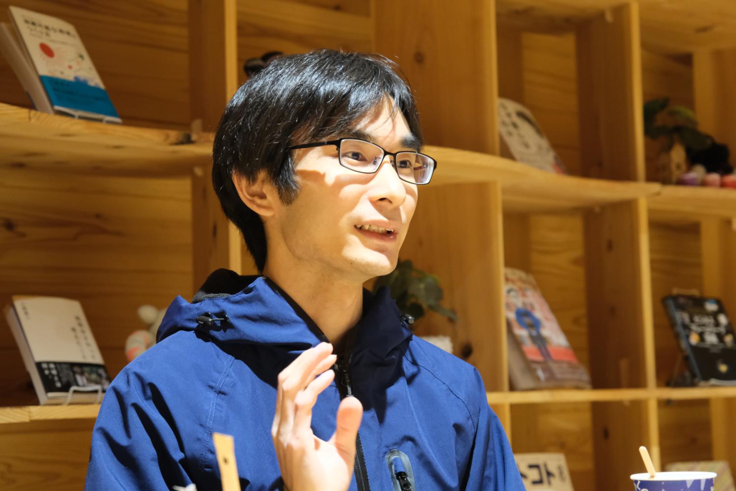 背景に棚があり、メガネをかけた菊池優さんが右手を上げ話をする様子の写真