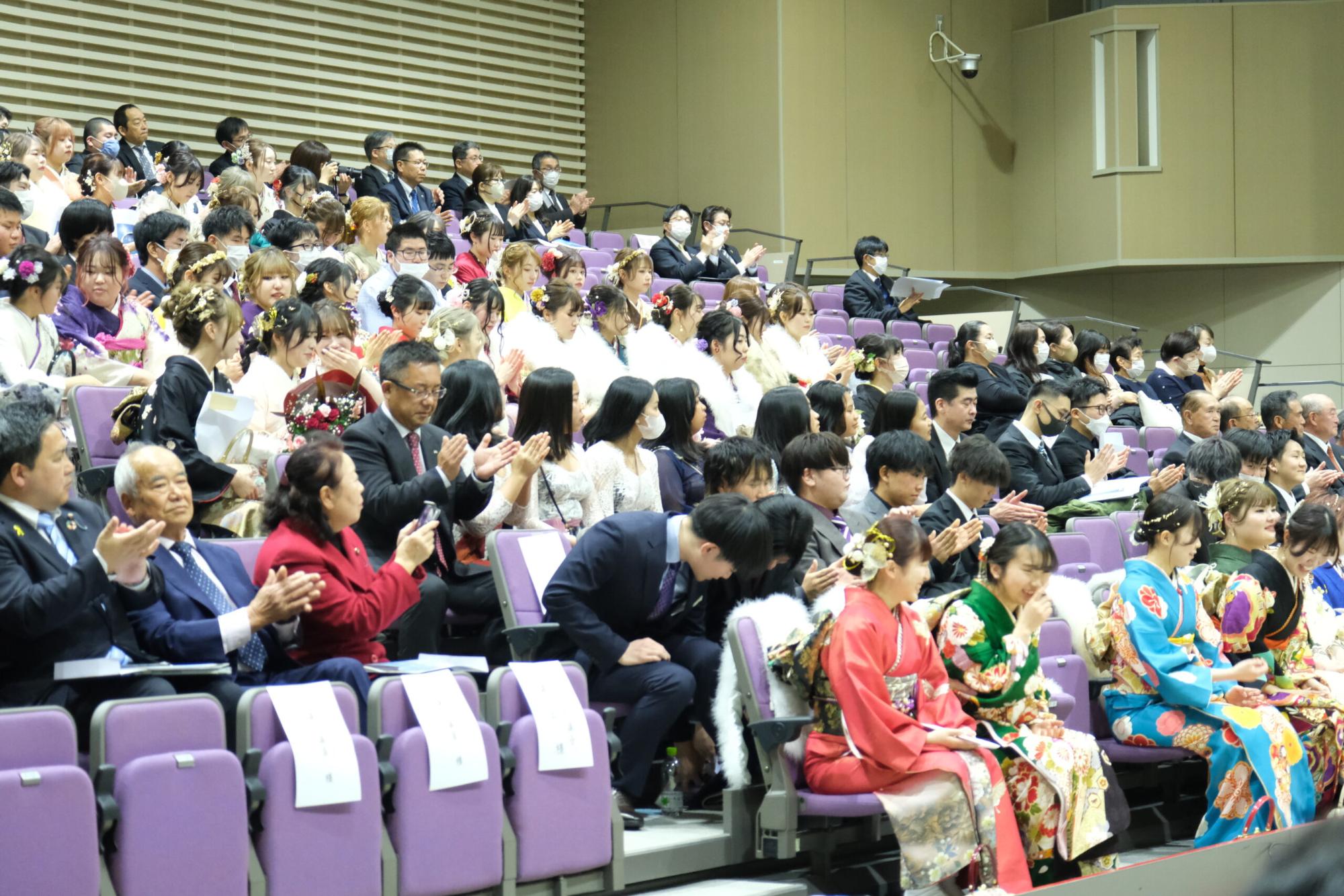 会場の客席で多くの新成人や来賓が拍手をしており、振袖やスーツを着た人々が座っている写真