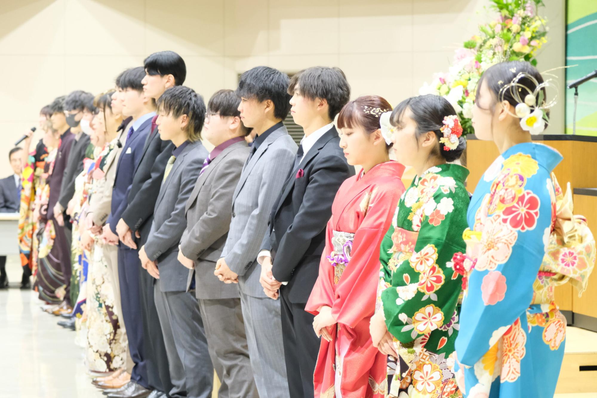 新成人の男女が正装や振袖を着て横一列に並び、前を向いて整列している写真