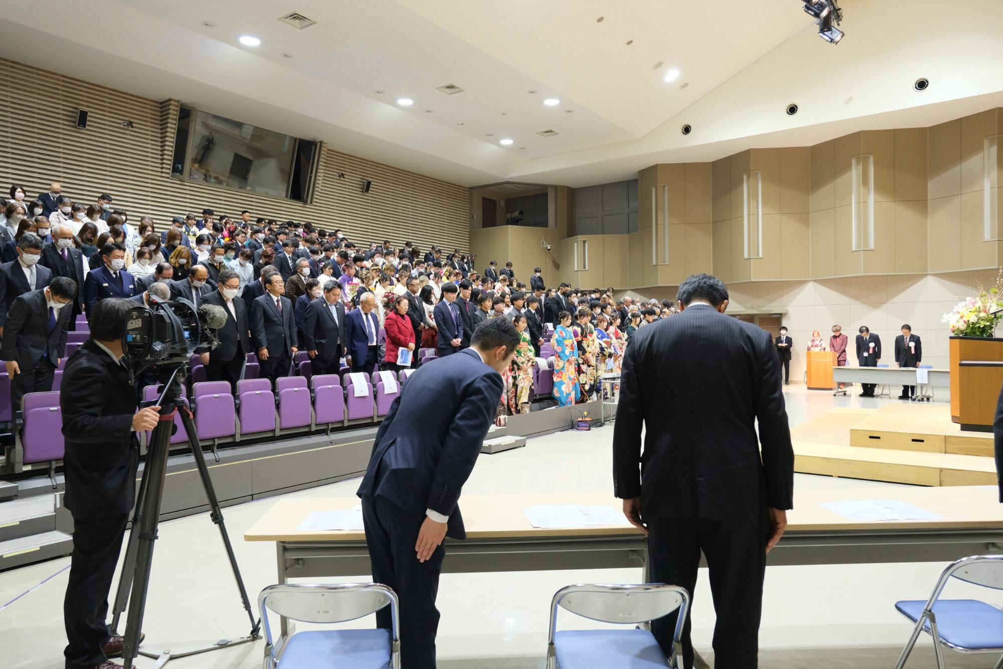 式典会場全体を広く捉えた構図で、壇上の主催者や来賓に向かって新成人や参列者たちが起立し、敬意を表している場面を写した、成人式中の一場面の写真
