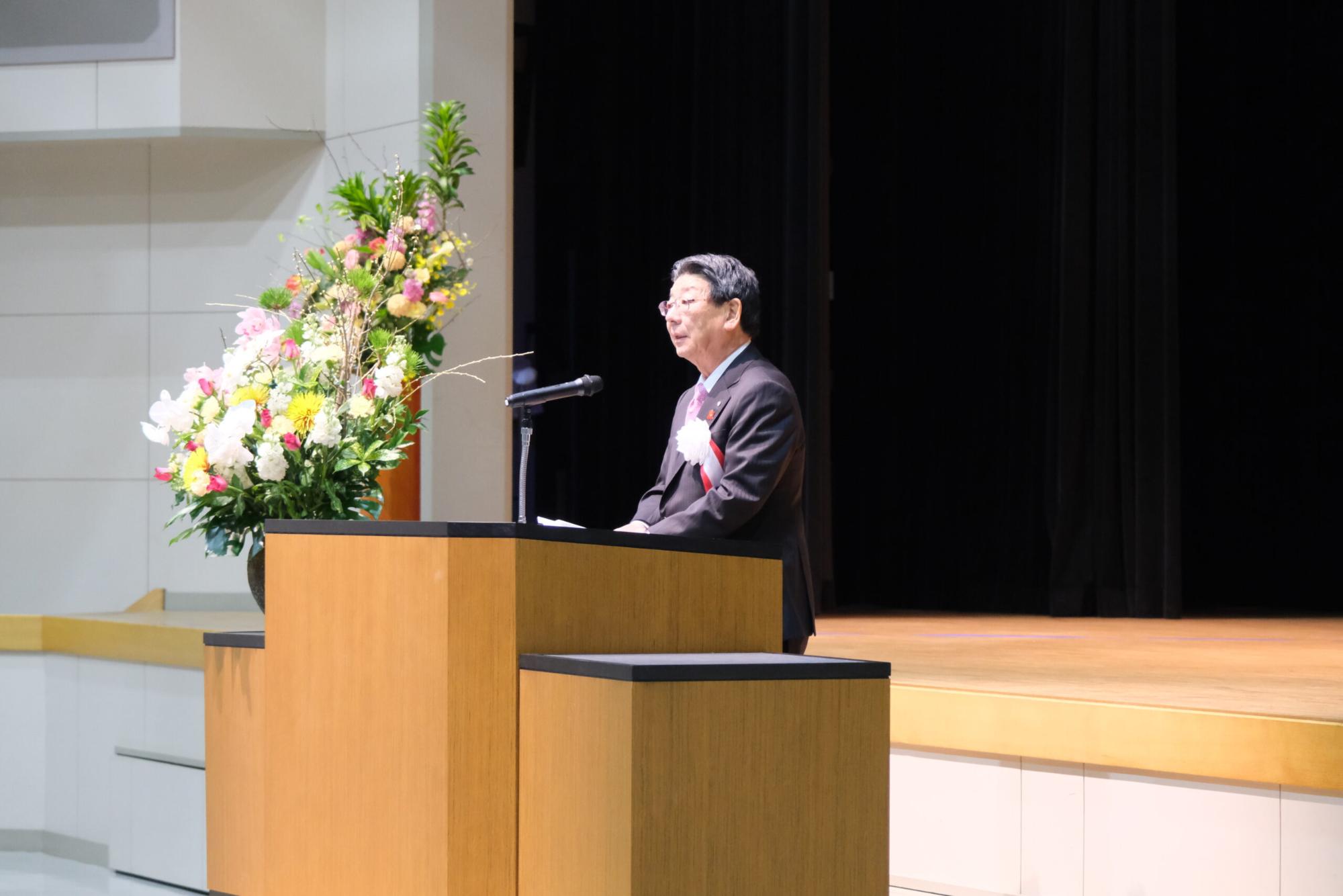 町長がステージ上の演台に立ち、来賓挨拶を述べており、横には大きな花が飾られている写真