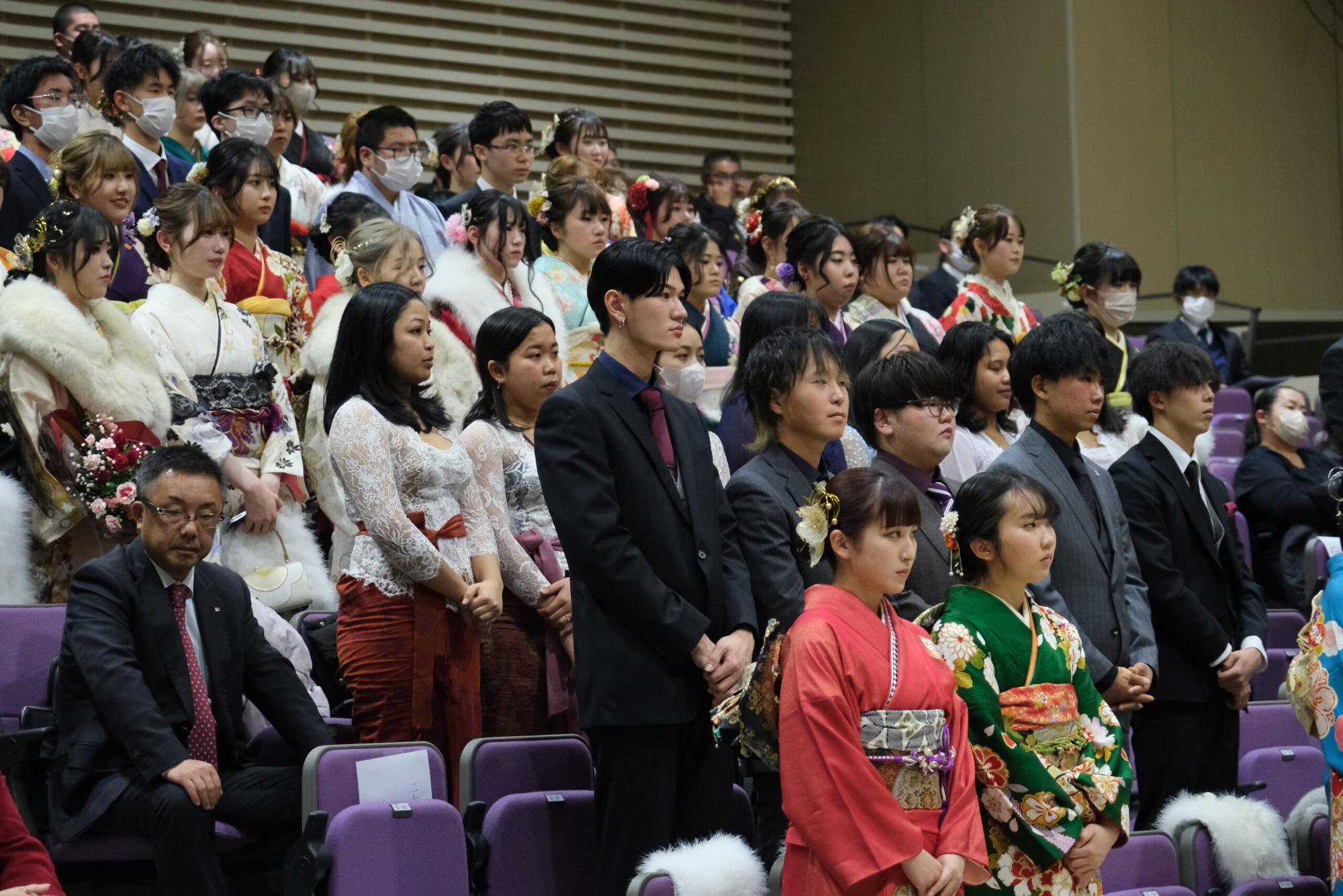 成人式の式典中に整列して立ち、真剣な表情で話を聞いている新成人たちの様子が写されており、色とりどりの振袖やスーツが並ぶ厳かな雰囲気の写真
