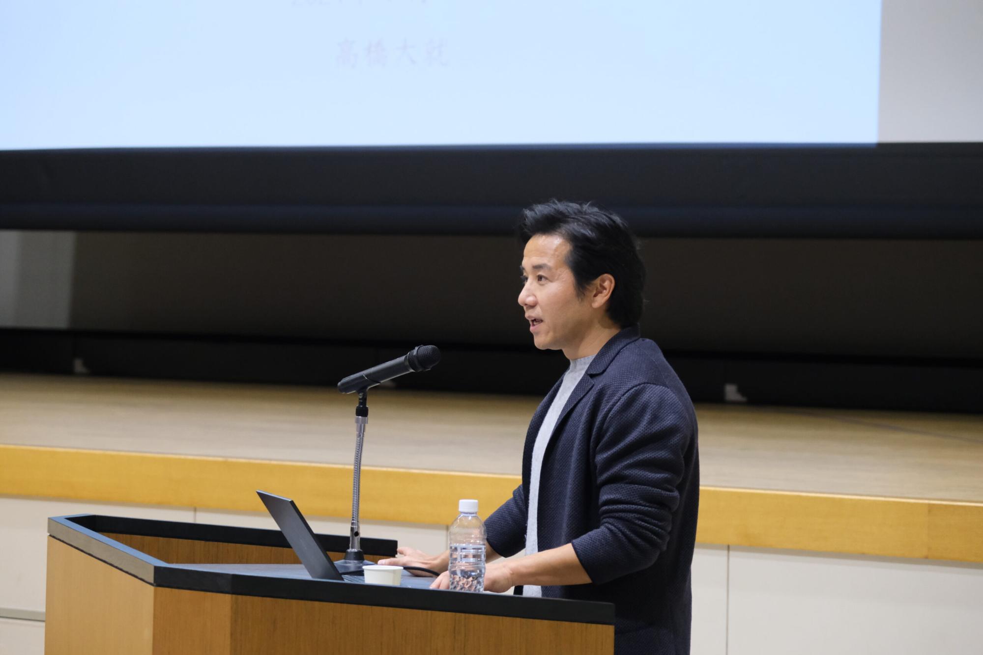 カジュアルな服装の講演者の男性が演台でパソコンとマイクを使って講演している写真