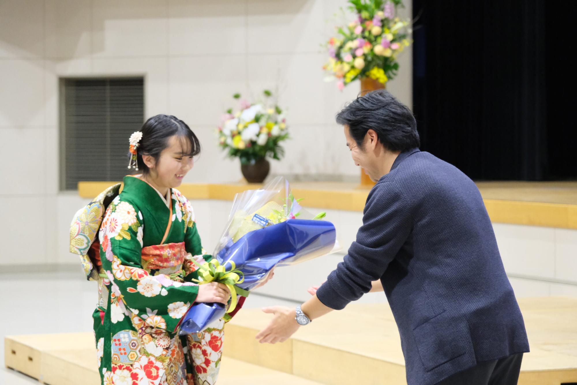 振袖姿の新成人の女性が壇上で講師の男性に花束を笑顔で手渡している写真