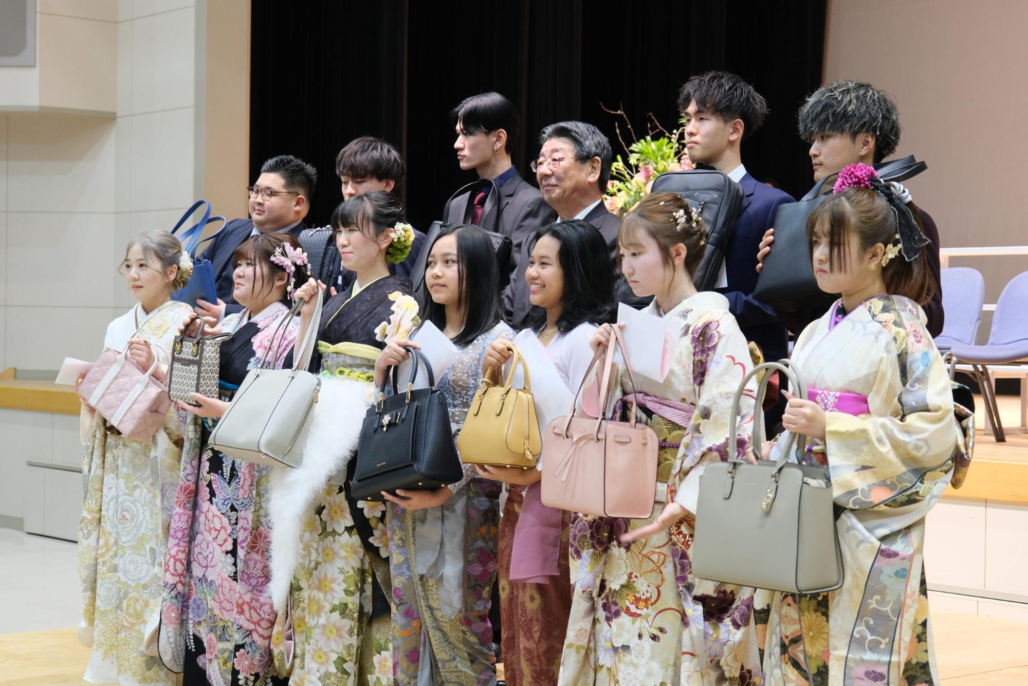 晴れ着やスーツを着た新成人たちが、バッグや記念品を手にして笑顔で記念撮影をしている、式典後の記念写真