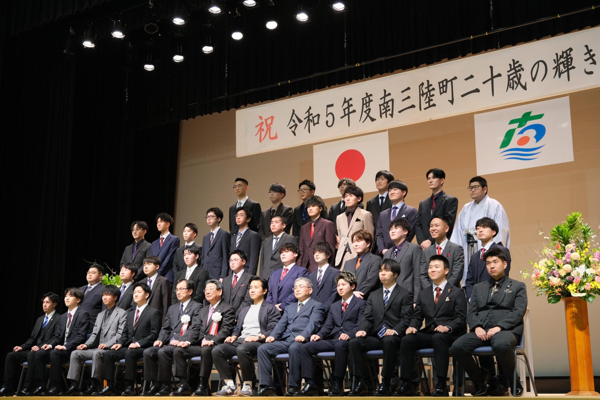 ステージ上でスーツ姿の新成人男性たちが整列し、町長や来賓らと一緒に国旗と町の旗を背に集合写真