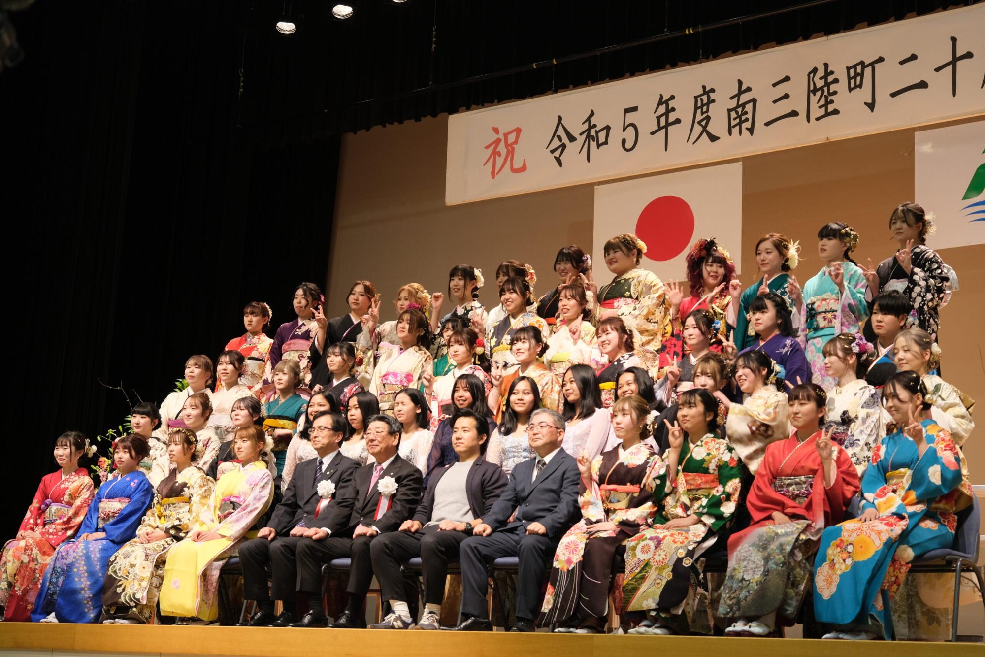 振袖姿の新成人女性たちがステージ上に数段に分かれて整列し、町長らとともに国旗と町の旗を背に記念撮影をしている、成人式での女性集合写真