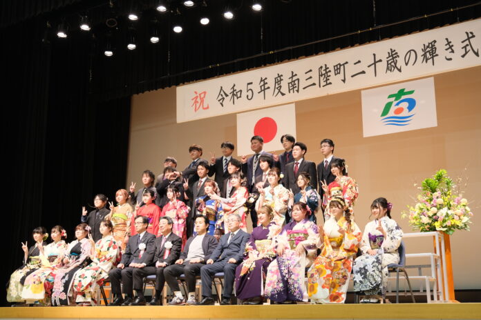 「令和5年度南三陸町二十歳の輝き式典」と書かれた垂れ幕の下で、振袖姿やスーツ姿の新成人たちと関係者が舞台上に並び、笑顔で記念写真を撮っている成人式の集合写真