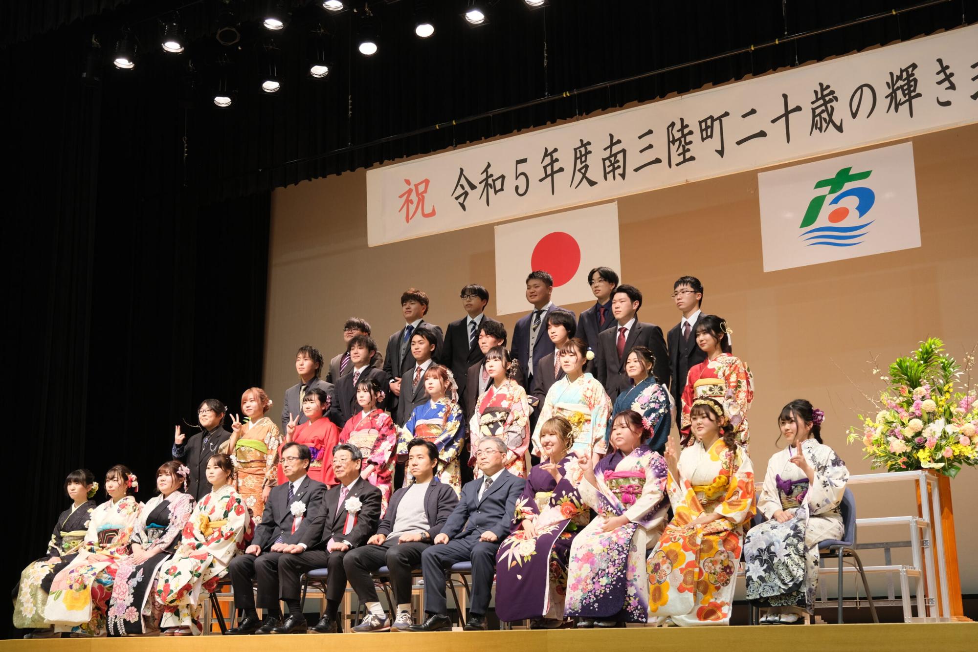 振袖姿の女性とスーツ姿の男性の新成人たちが混合でステージ上に整列し、町長や来賓らと共に記念撮影をしている、男女合同の成人式集合写真