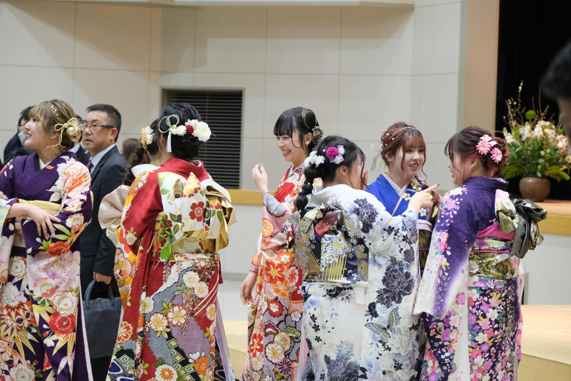 振袖姿の新成人女性たちが式典会場内で談笑しながら再会を喜び合い、それぞれの着物や髪飾りが華やかに映えている、成人式当日の交流風景の写真