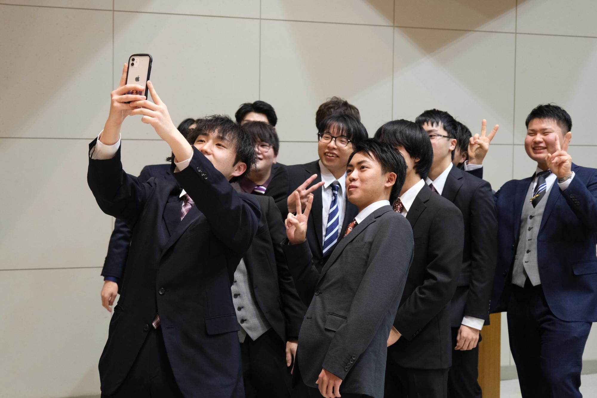 スーツを着た新成人の男性たちがスマートフォンで記念撮影をしており、皆が笑顔でピースサインをしながら楽しい雰囲気を共有している、成人式会場での写真
