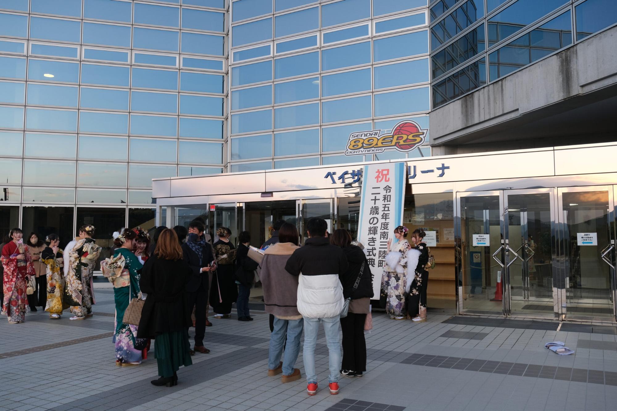 ガラス張りの「ベイサイドアリーナ」正面入口の前に振袖姿の新成人や家族・知人が集まり、成人式の立て看板の前で記念撮影や会話を楽しんでいる、式典直前の様子を捉えた写真
