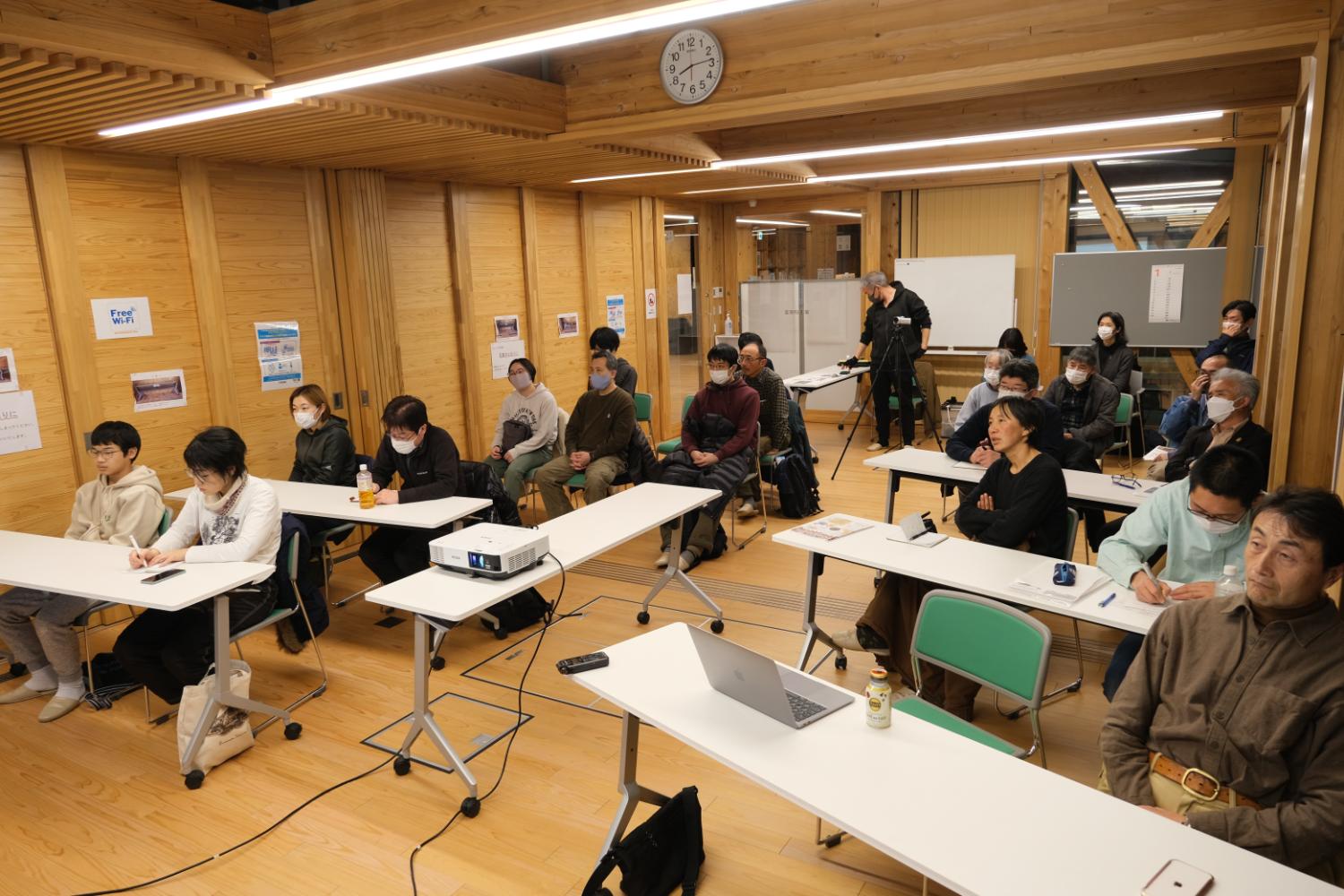 机と椅子が並ぶ室内に、マスクした地域住民・参加者が前方スクリーンに見入りながらノートを取るなどして真剣に聴講している町民向け講座の写真