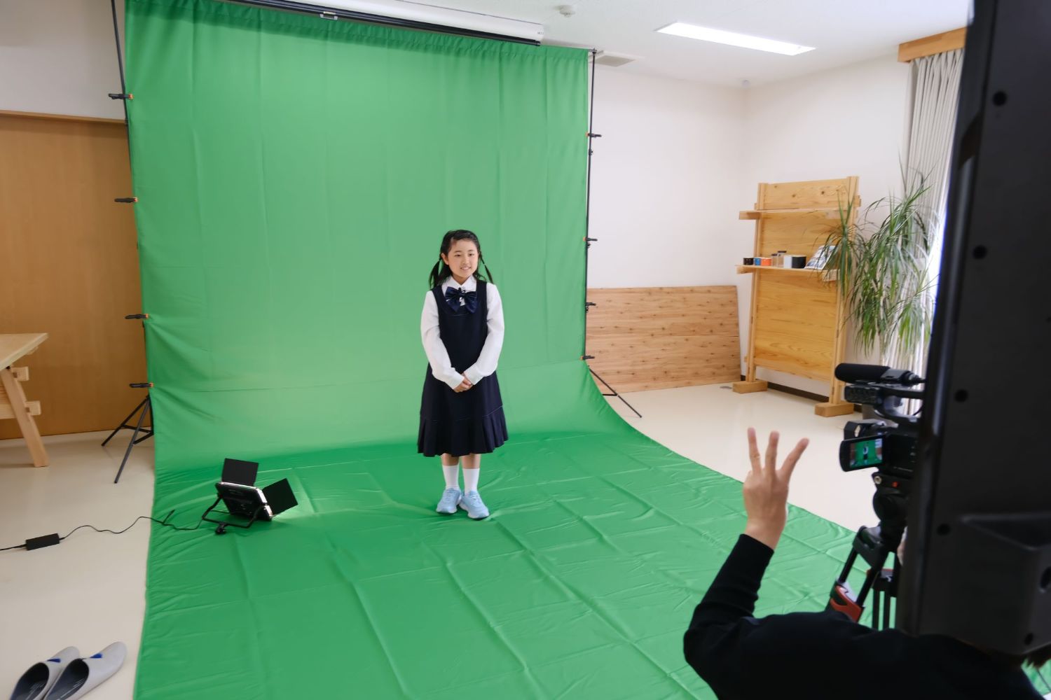 白いシャツに紺色のワンピースを着た女子児童が緑色の布を背景に撮影をしている写真