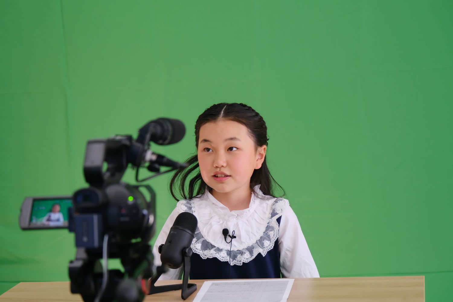 緑のクロマキーの前でリポーターのあこさんが机の前に座り、マイク付きのカメラの前でニュース原稿を読み上げている様子の写真